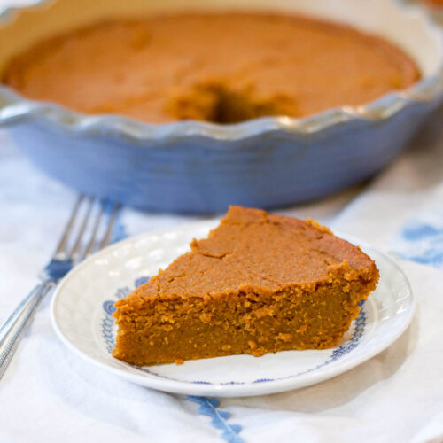 A slice of crustless pumpkin pie on a small plate sits in front of a pie dish with the remaining pie, accompanied by a fork beside the plate.