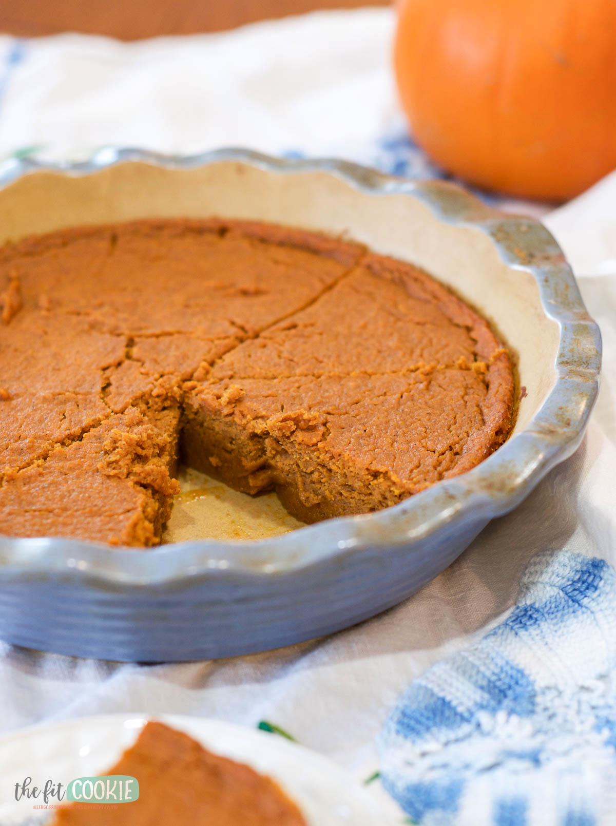 A crustless pumpkin pie, partially sliced in a ceramic pie dish, rests on a white cloth with a pumpkin in the background.