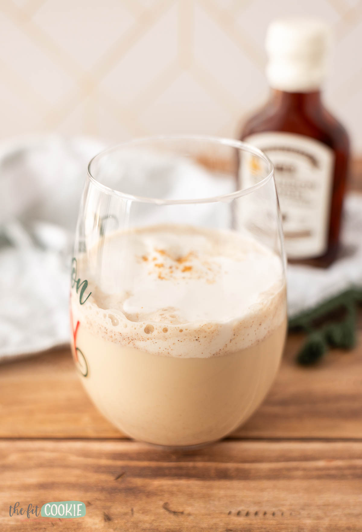 A glass of frothy dairy free egg nog topped with a sprinkle of spice, placed on a wooden surface with a bottle and cloth in the background.