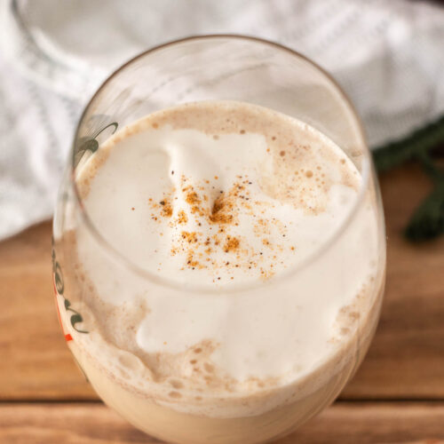 A glass of creamy, light brown dairy free egg nog topped with foam and a sprinkle of ground spice, placed on a wooden surface.