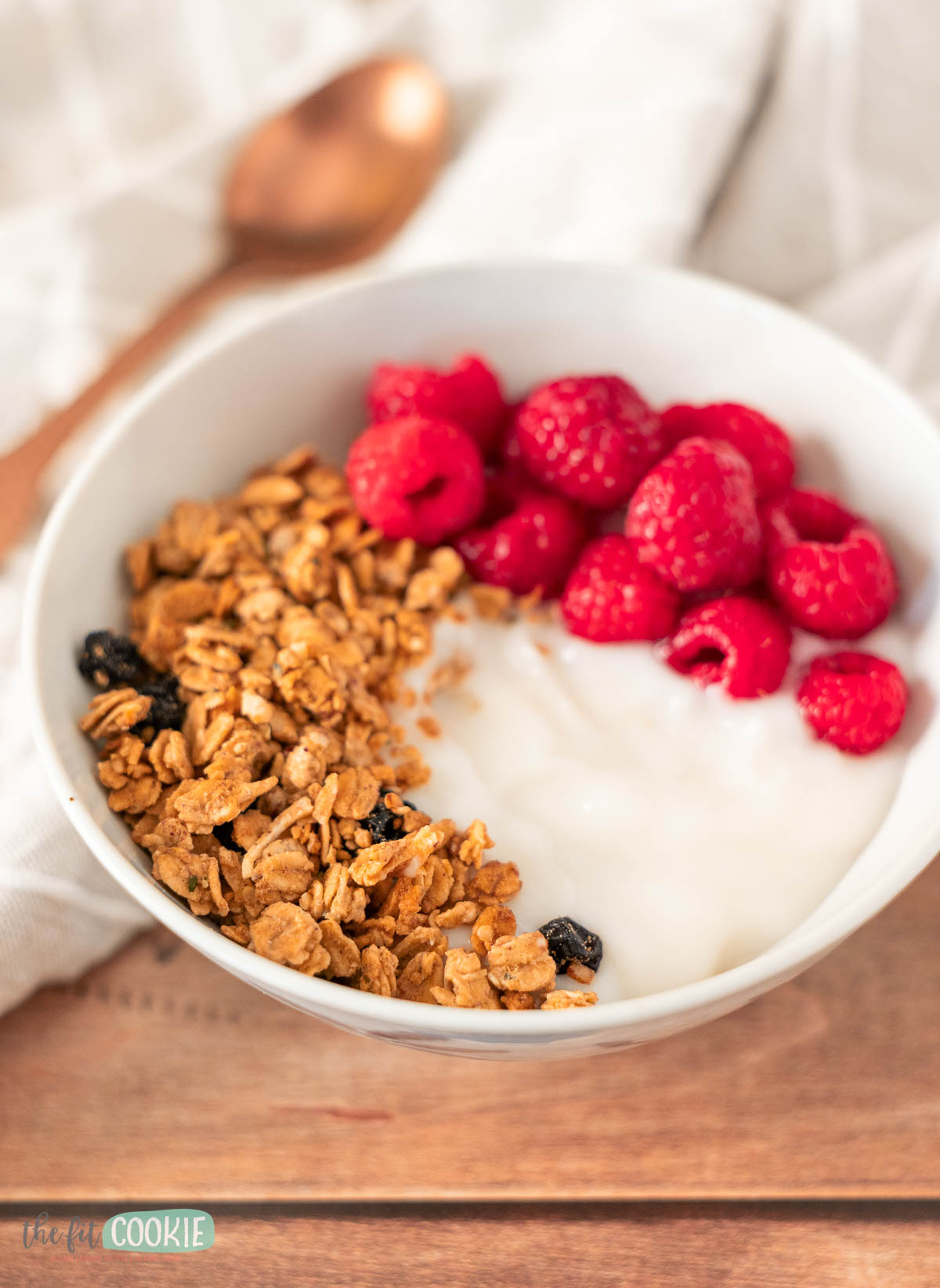 A white bowl filled with yogurt, maple sunbutter granola, and fresh raspberries sits on a wooden surface.