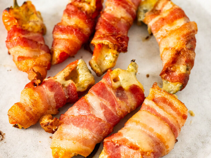 Six bacon-wrapped jalapeño poppers, reminiscent of stuffed baby peppers, filled with melted cheese are arranged on a round white plate.
