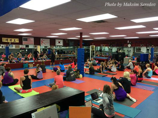A large group of people sits on mats in a martial arts studio, listening to an instructor at the front of the room. Martial arts equipment, PiYo mats, and flags are visible on the walls.