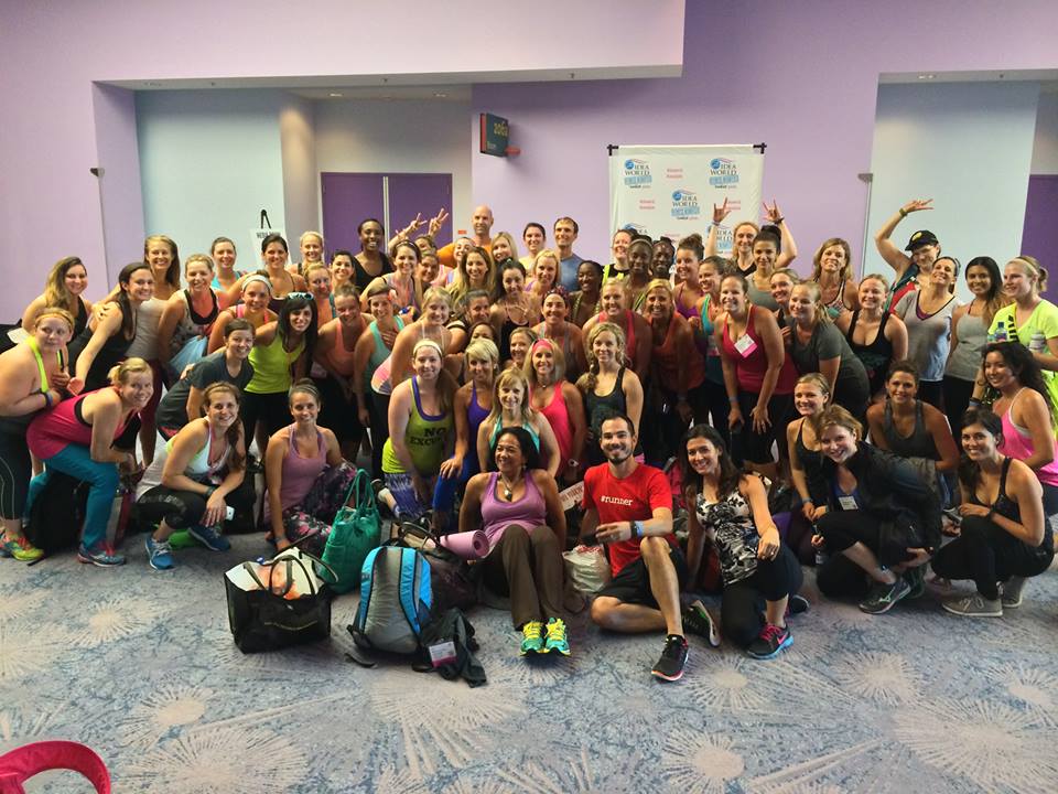 Large group of people in athletic wear pose and smile together indoors after a PiYo class, some kneeling in front, with gym bags visible on the floor.