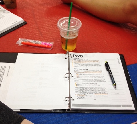 An open binder with papers labeled "PiYo," a black pen, a highlighter, a plastic cup with a drink, and a wrapped snack are arranged on a red and blue surface.
