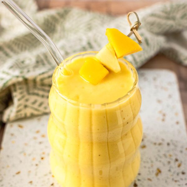 A glass of yellow tropical smoothie with a glass straw, garnished with mango and pineapple pieces, sits on a speckled white surface with a green patterned cloth in the background.