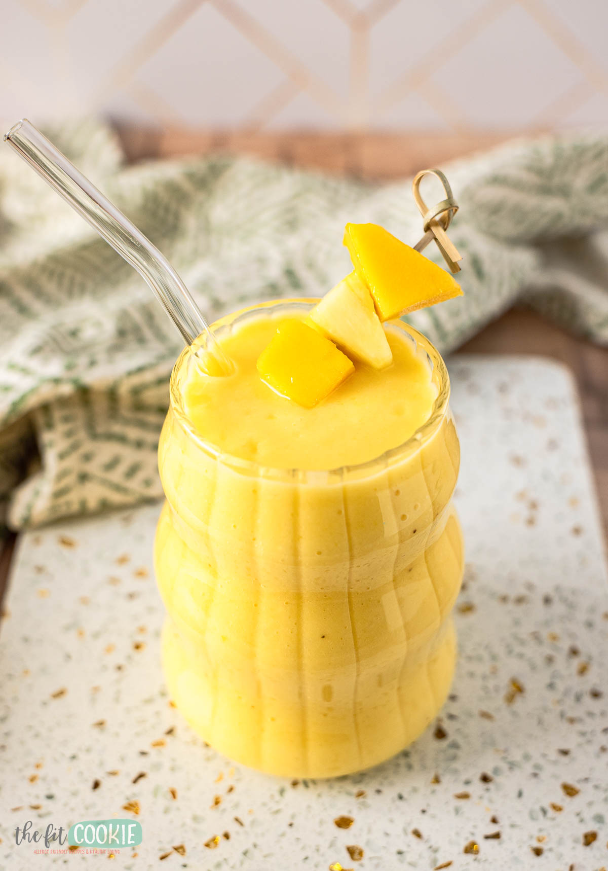 A glass of yellow tropical smoothie with a glass straw, garnished with mango and pineapple pieces, sits on a speckled white surface with a green patterned cloth in the background.