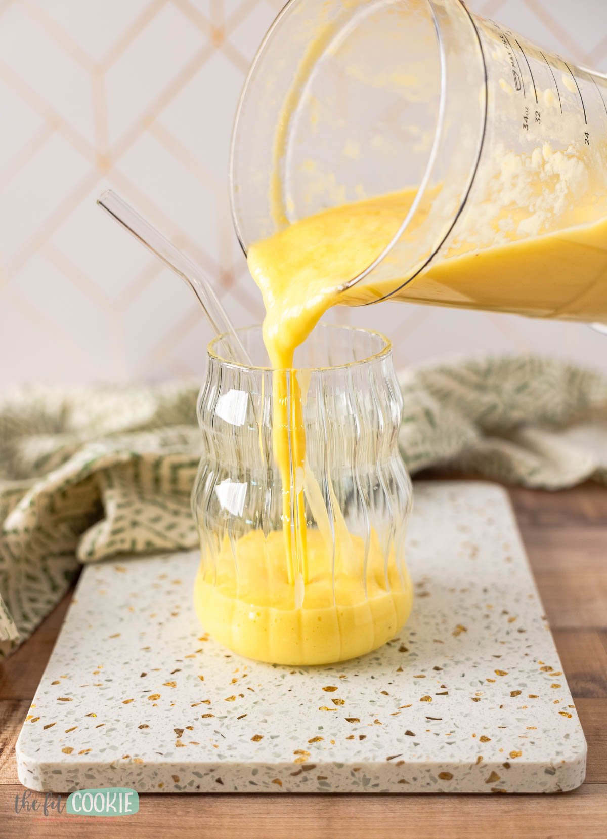 A thick yellow tropical smoothie is being poured from a blender into a wavy glass with a clear straw, set on a terrazzo board.