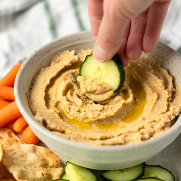 A hand dips a slice of cucumber into a bowl of cauliflower hummus, surrounded by carrot sticks, crackers, and cucumber slices.