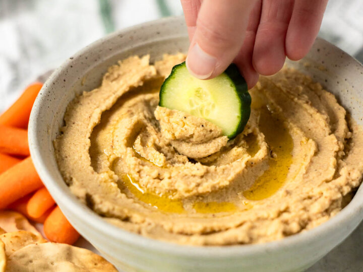 A hand dips a slice of cucumber into a bowl of cauliflower hummus, surrounded by carrot sticks, crackers, and cucumber slices.