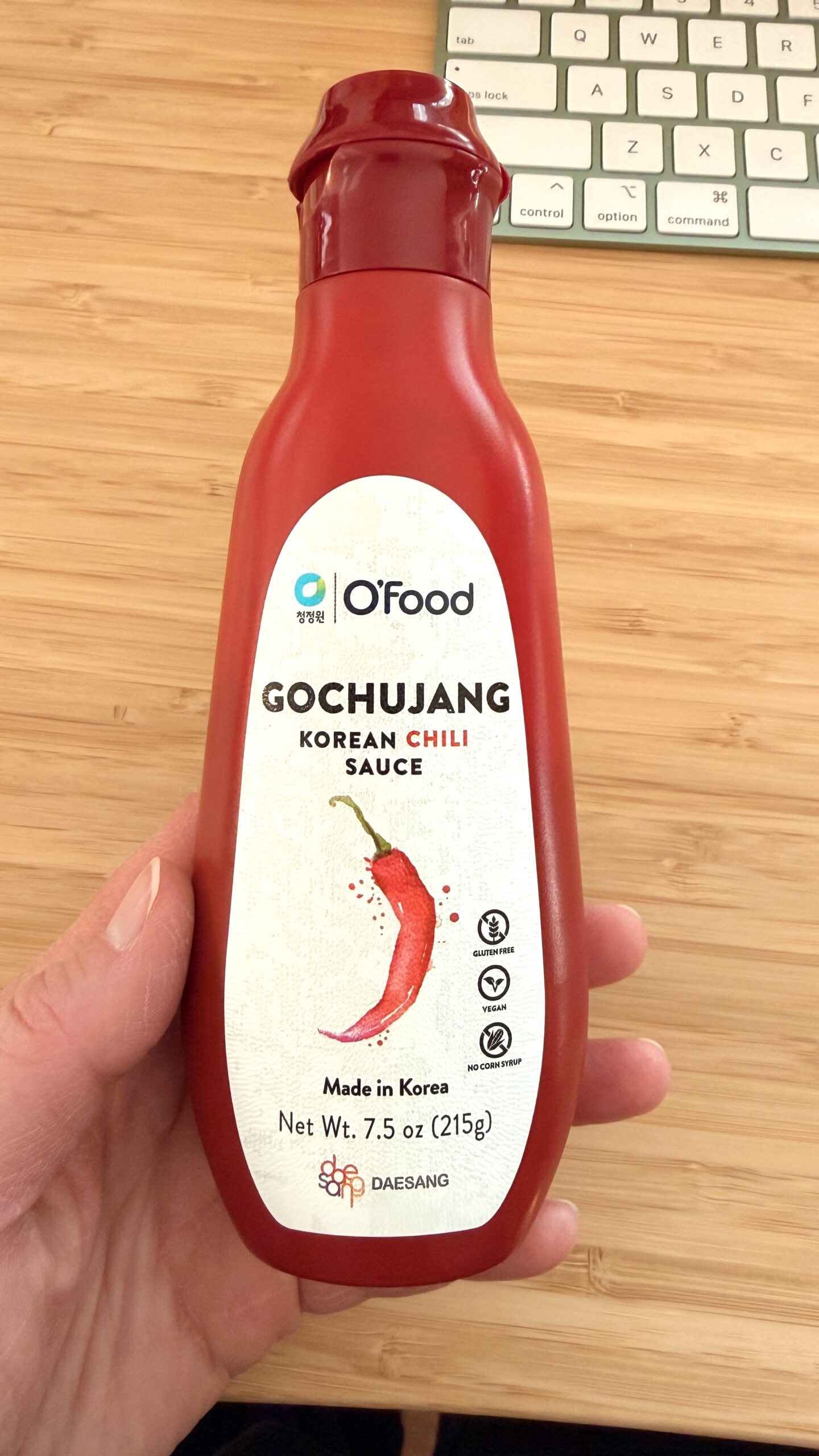 A hand holds a red bottle of O'Food Gochujang Korean Chili Sauce, 7.5 oz (215g), perfect for soy free cooking. The label features a chili pepper graphic, with a keyboard visible in the background.