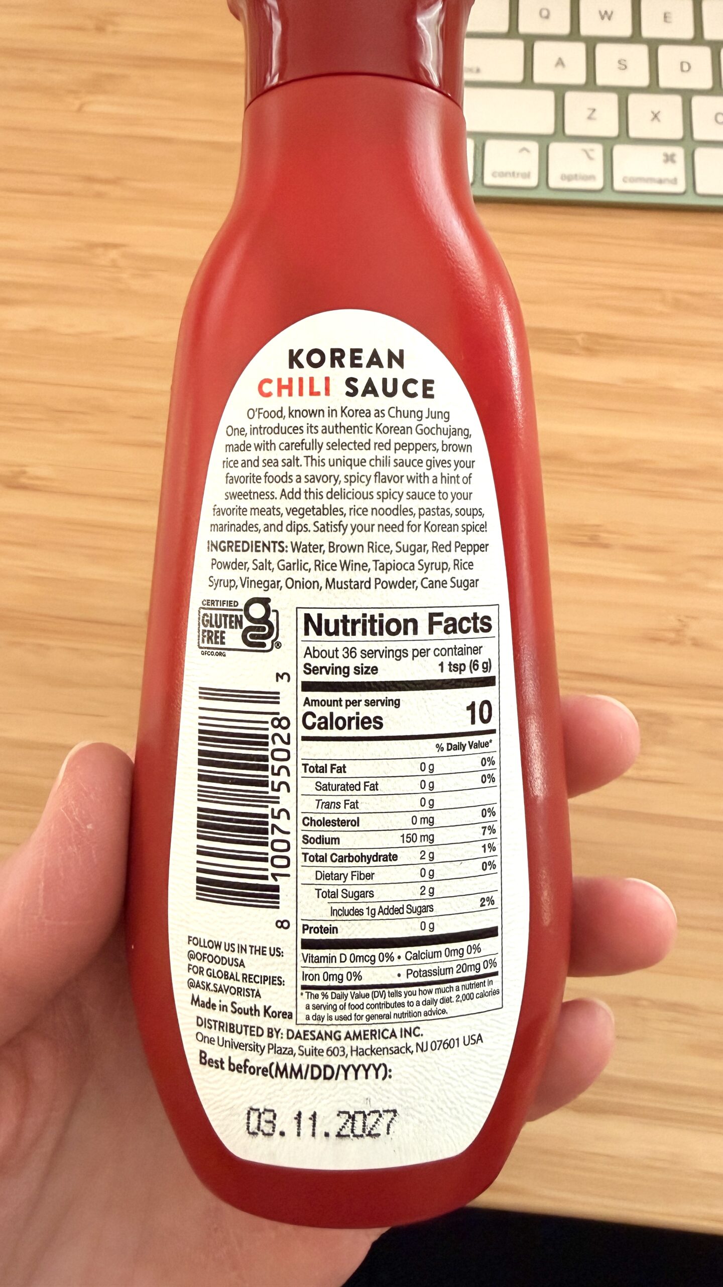A hand holds a bottle of soy free Korean chili sauce, showing nutrition facts, ingredients, a barcode, and an expiration date of 03.11.2027 on the back label. A keyboard is in the background—perfect for soy free cooking enthusiasts.