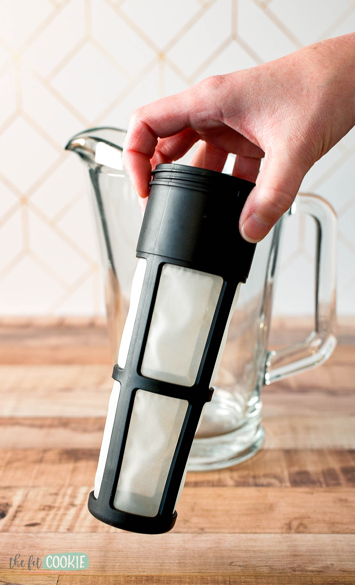 A hand holds a reusable mesh filter for cold brew coffee above an empty glass pitcher on a wooden surface.