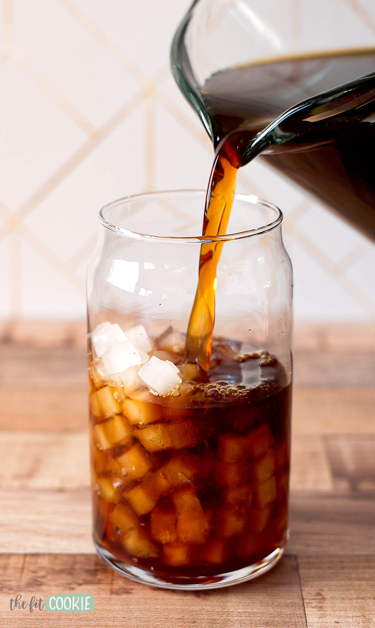 A glass filled with ice cubes is being filled with cold brew coffee, from a pitcher.
