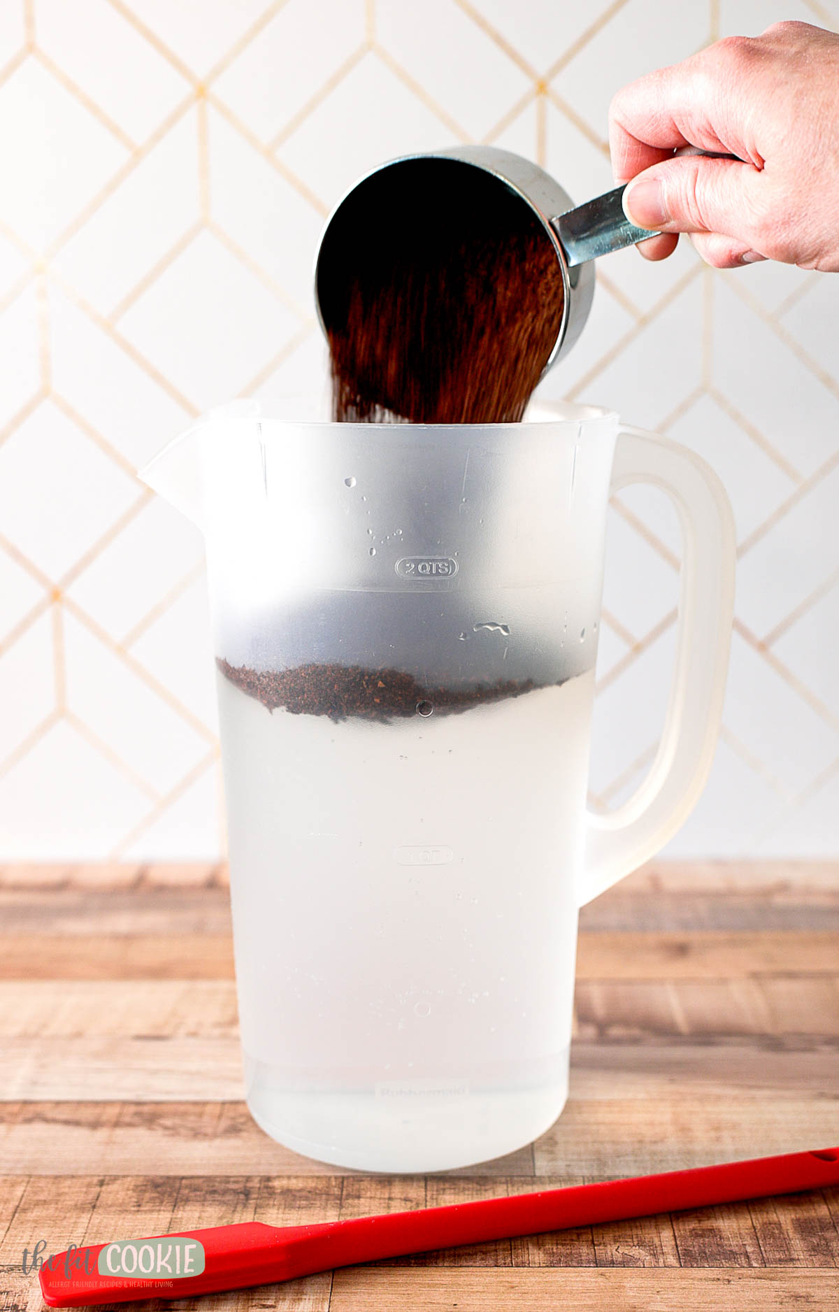 A hand pours ground coffee for cold brew coffee from a measuring cup into a plastic pitcher filled with water.