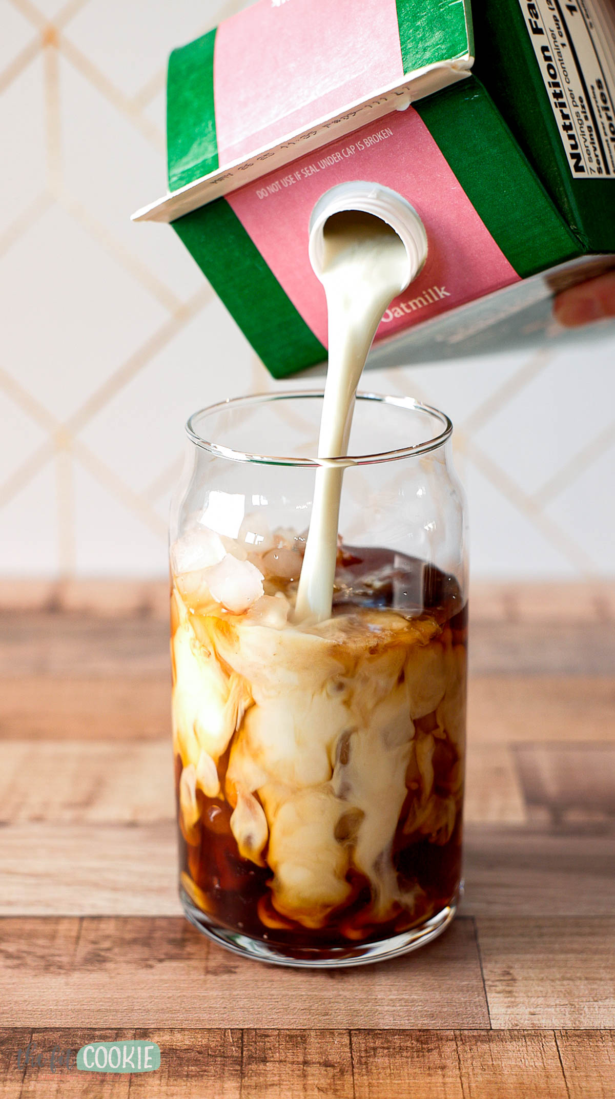 Oat milk being poured from a carton into a glass of cold brew coffee on a wooden countertop.