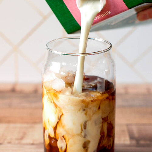 Oat milk is being poured from a carton into a glass of cold brew coffee on a wooden surface.