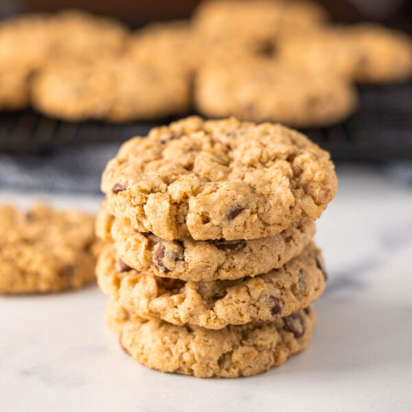 A stack of four oatmeal chocolate chip cookies sits on a white surface, with more freshly baked oatmeal chocolate chip cookies cooling on a rack in the blurred background.
