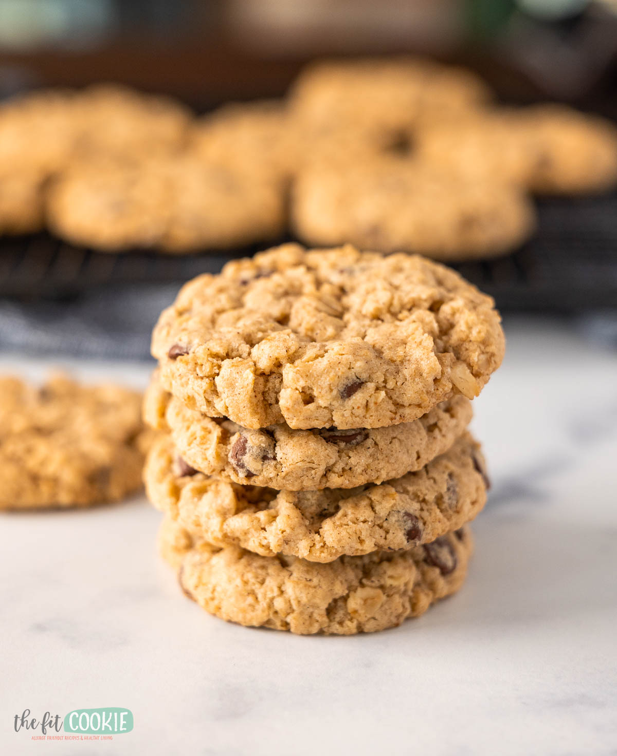 A stack of four oatmeal chocolate chip cookies sits on a white surface, with more freshly baked oatmeal chocolate chip cookies cooling on a rack in the blurred background.