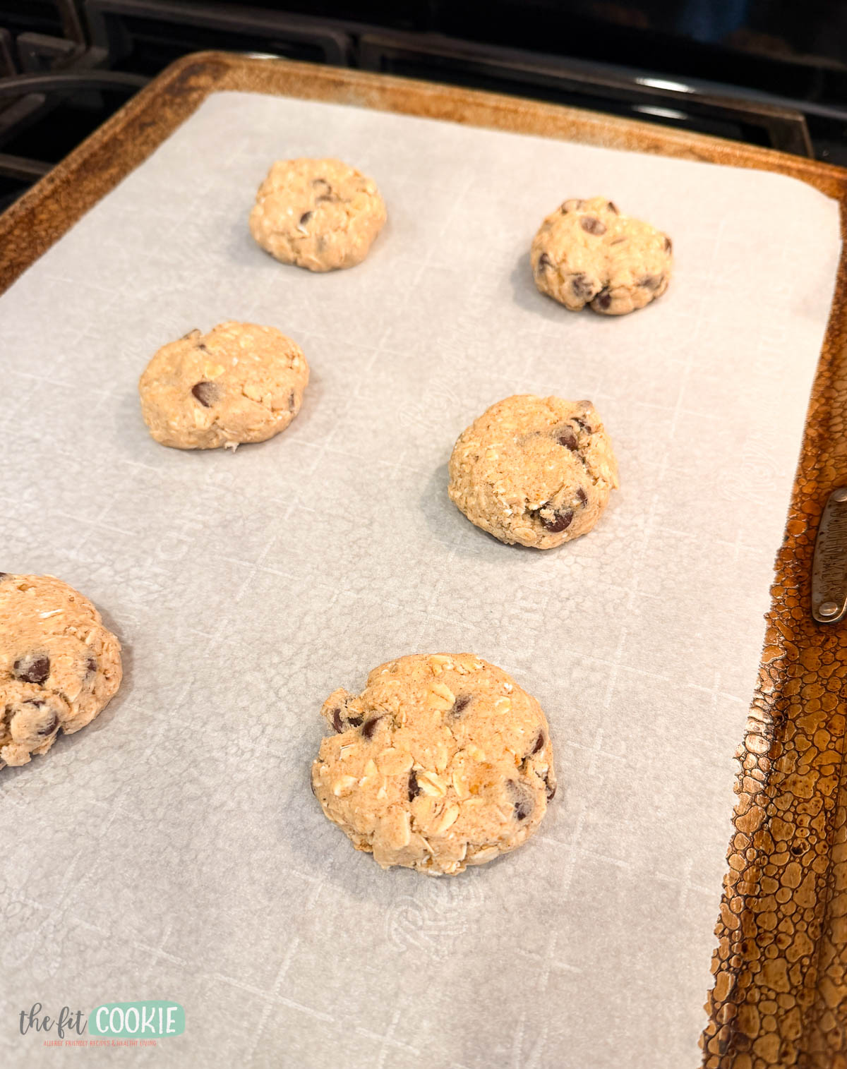 Six unbaked oatmeal chocolate chip cookies dough balls on a parchment-lined baking sheet.