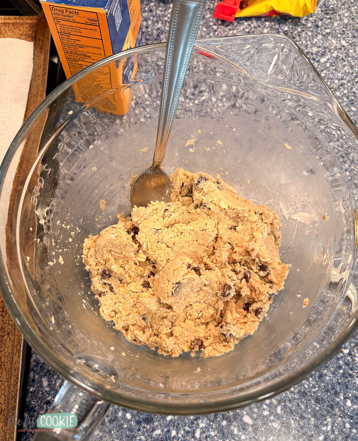 A glass mixing bowl with oatmeal chocolate chip cookie dough and a metal spoon sits on a blue speckled countertop.