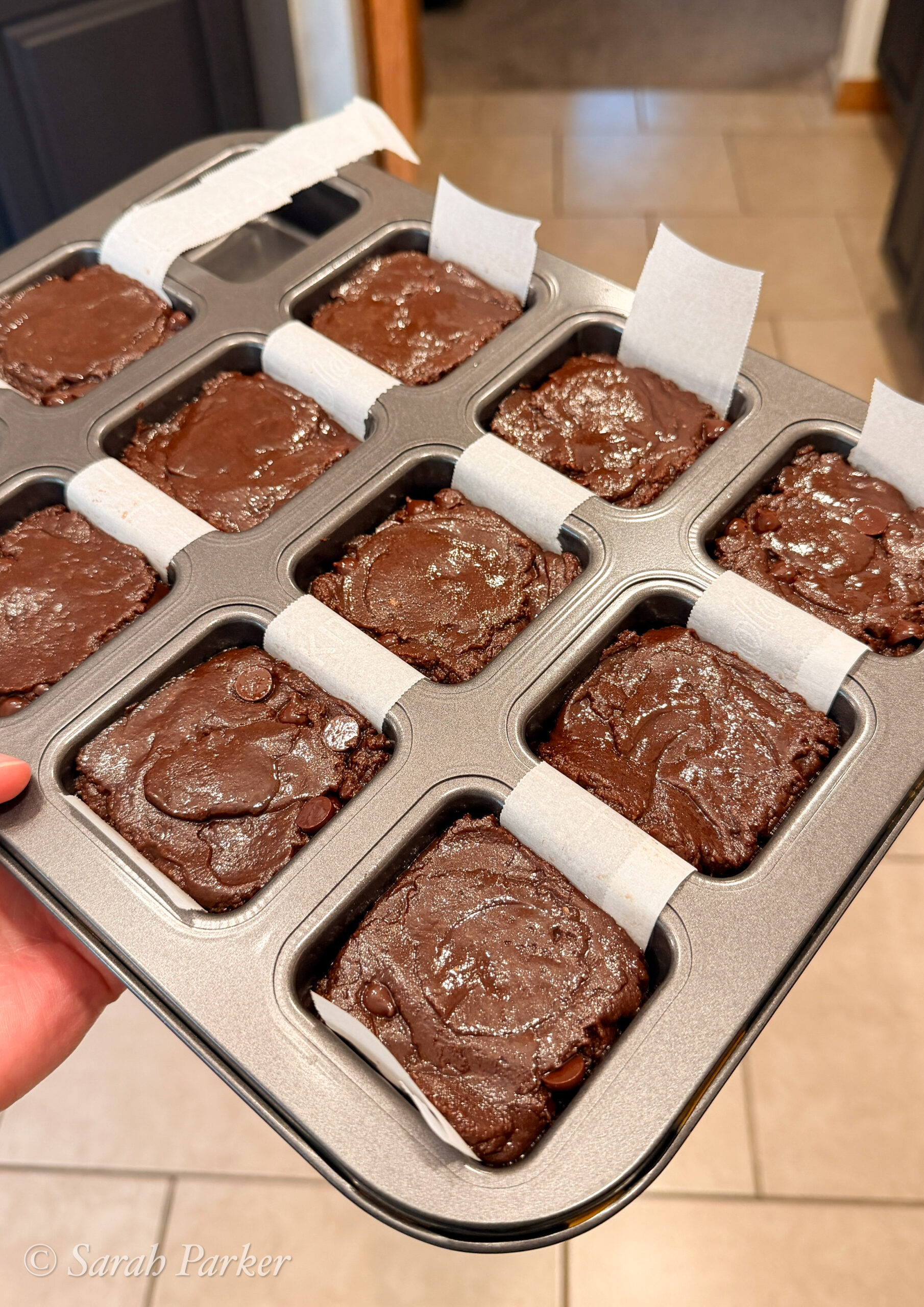 A hand holds a baking tray of unbaked Fudge Brownies in twelve square molds, each lined with parchment paper strips.