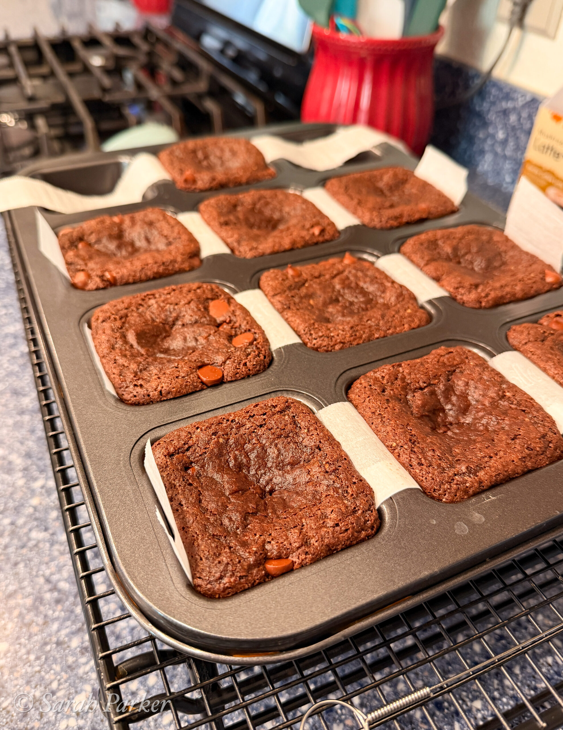 A divided brownie pan filled with freshly baked square fudge brownies, each resting on parchment strips, is cooling on a wire rack in a kitchen.