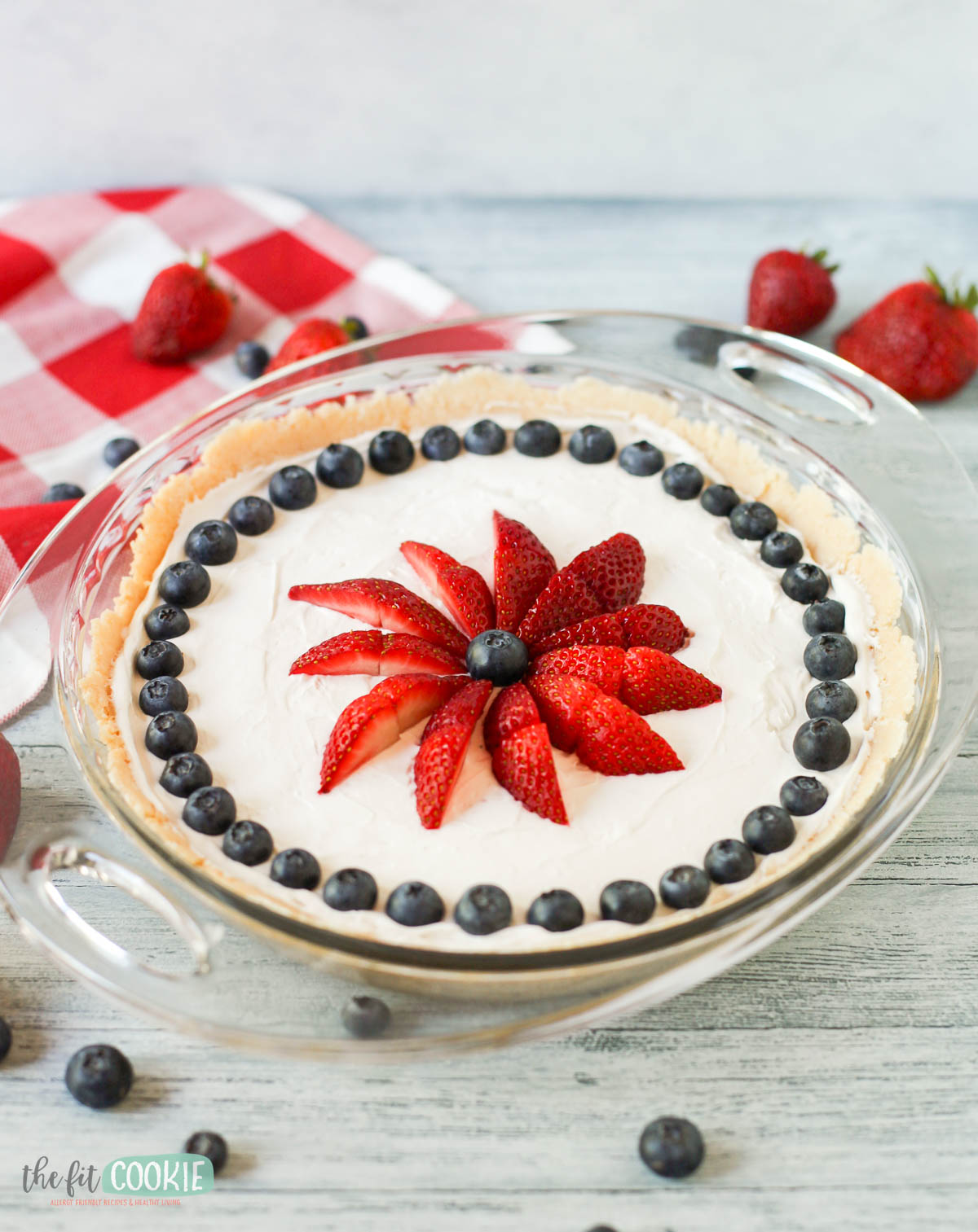 A dairy free vanilla cheesecake topped with halved strawberries arranged in a flower shape and a ring of blueberries, served in a glass pie dish on a light wooden surface.