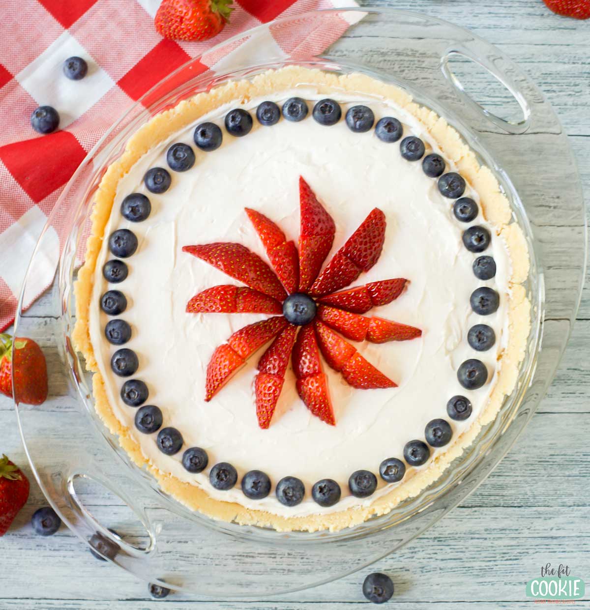 A dairy free vanilla cheesecake with a creamy filling, topped with a ring of blueberries and a starburst pattern of sliced strawberries, sits on a wooden table with a red checkered cloth and scattered berries.