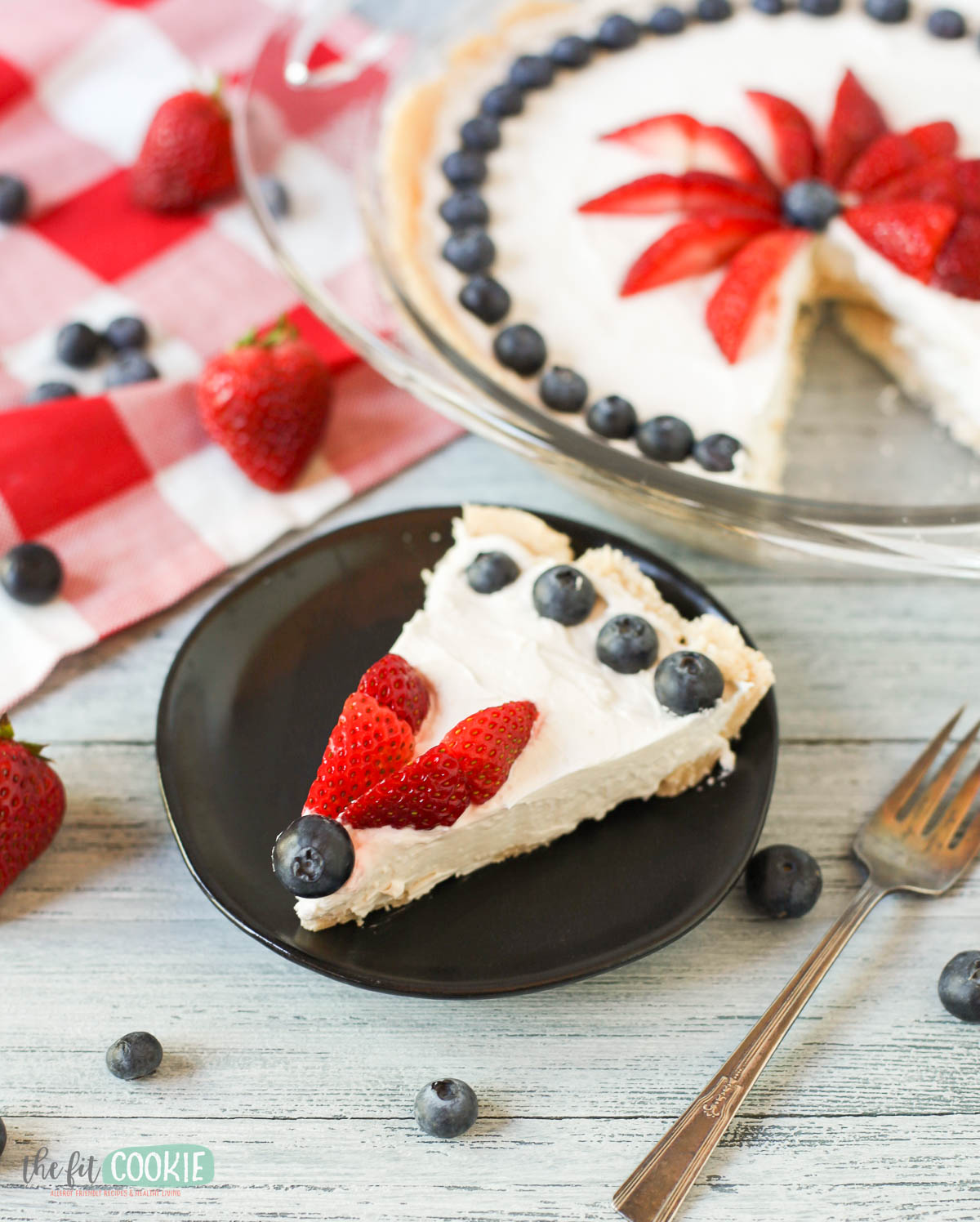 A slice of dairy free vanilla cheesecake on a black plate, decorated with sliced strawberries and blueberries, sits next to a whole pie and fresh berries on the table.