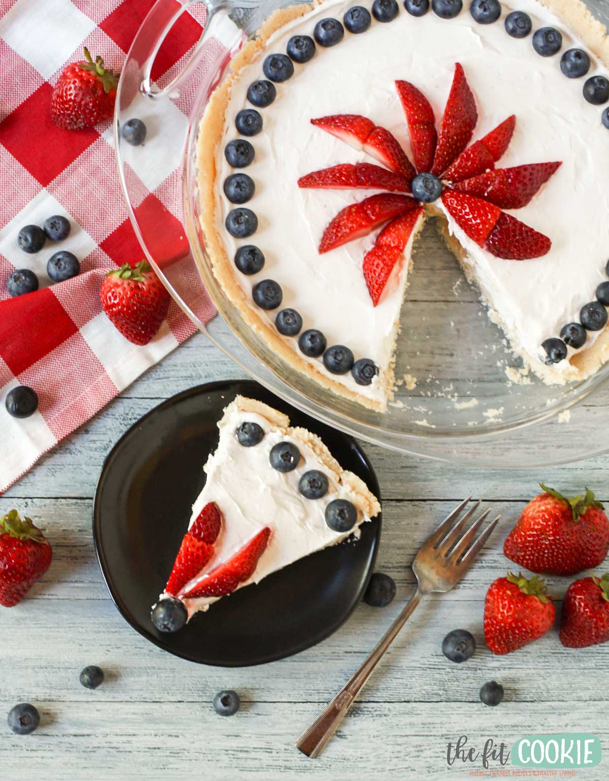 A dairy free vanilla cheesecake with a creamy white topping, decorated with strawberries and blueberries, sits next to a slice on a black plate, on a wooden table with scattered berries and a red checked cloth.