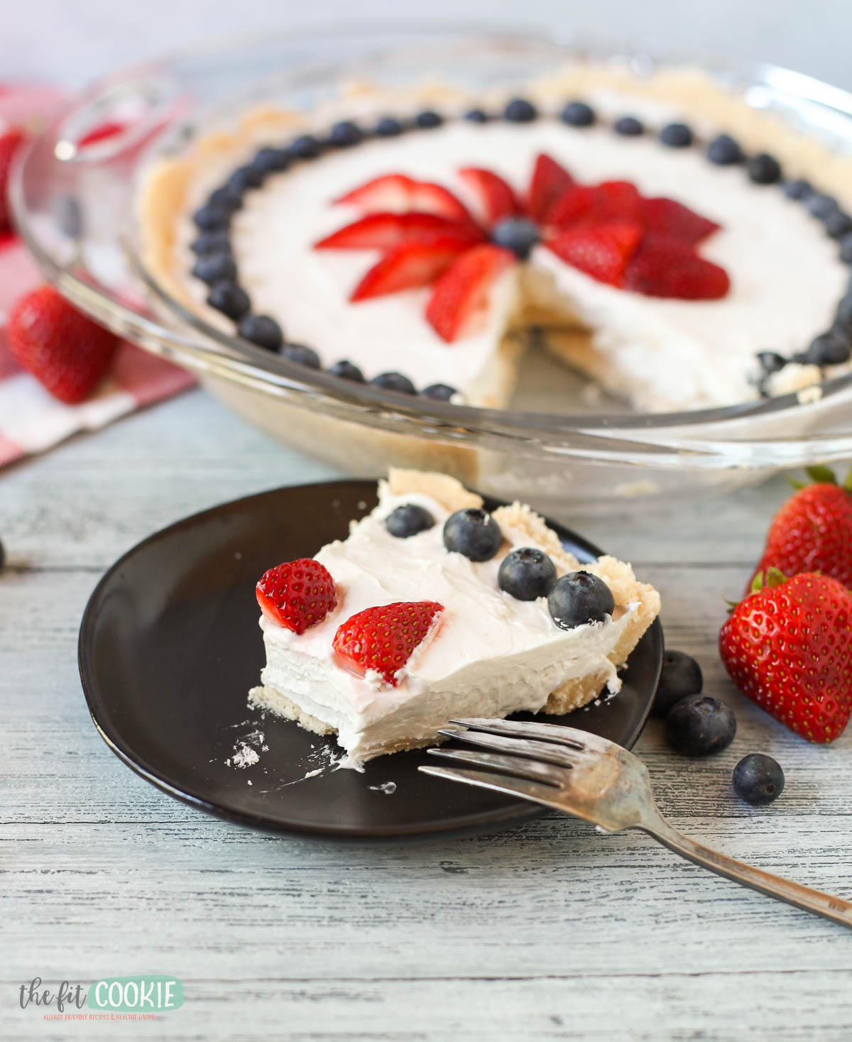 A slice of dairy free vanilla cheesecake topped with strawberries and blueberries on a black plate, with a fork and the remaining pie in the background.