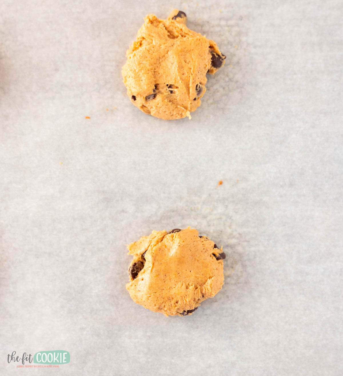 Two scoops of pumpkin chocolate chip cookie dough are placed on a sheet of parchment paper, ready for baking.