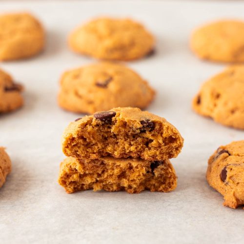 A pumpkin chocolate chip cookie broken in half sits on parchment paper, surrounded by several whole cookies in the background.