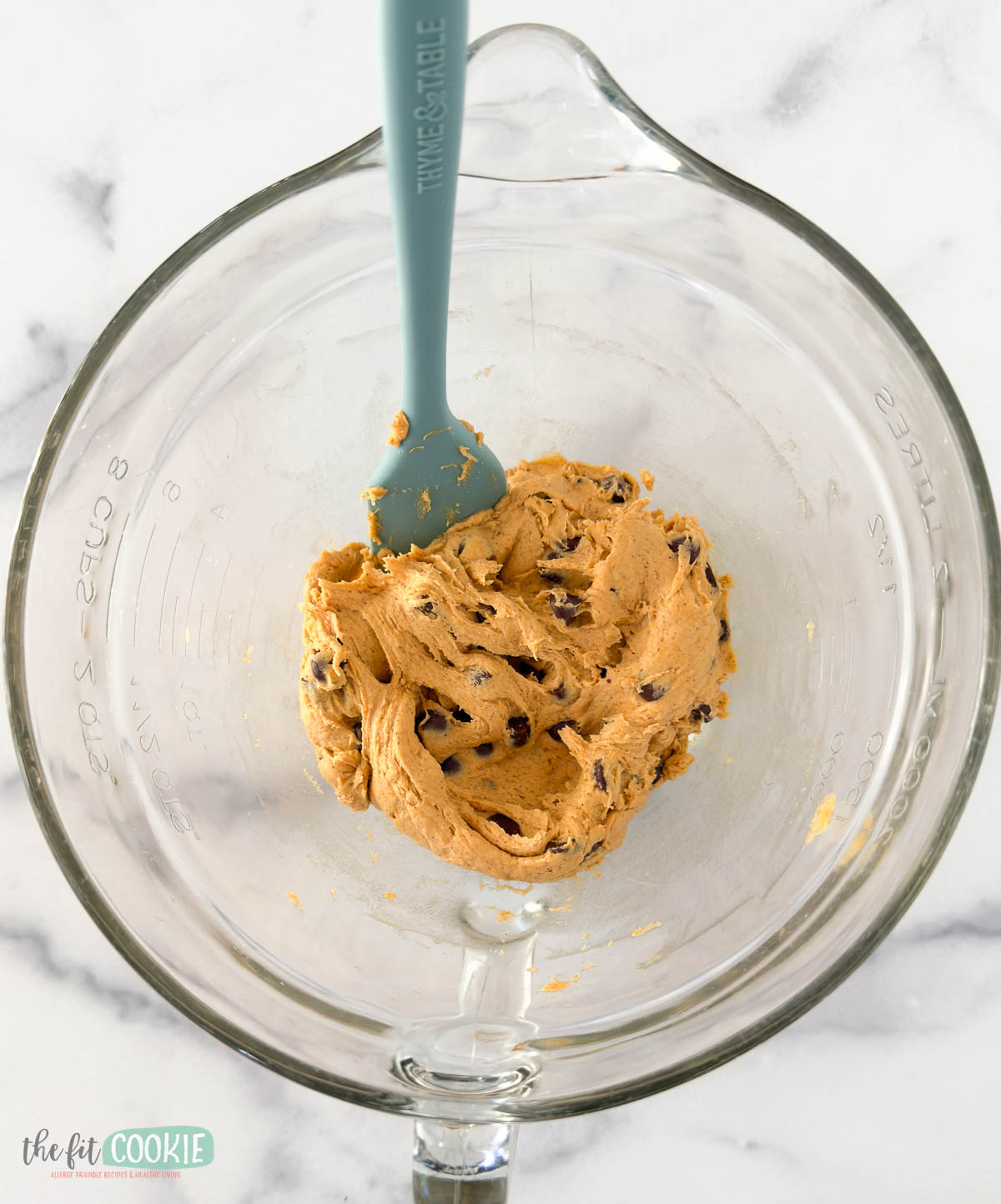 A glass mixing bowl containing pumpkin chocolate chip cookie dough and a blue spatula on a white marble surface.