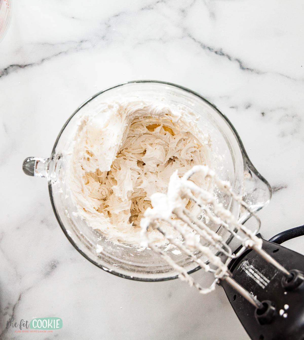 Electric hand mixer with beaters rests in a glass bowl filled with dairy free buttercream frosting on a white marble countertop.