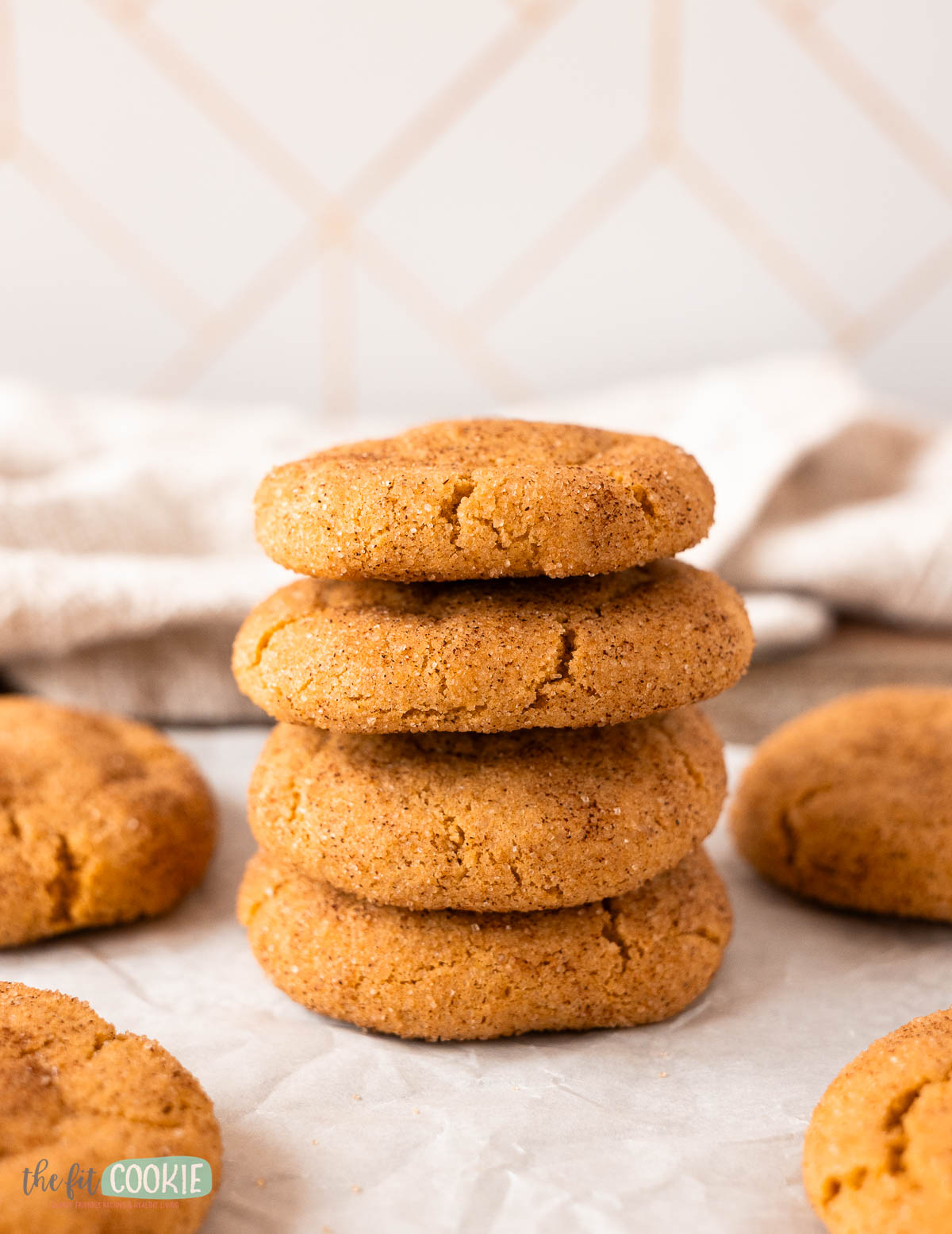A stack of five round, golden-brown pumpkin snickerdoodles is placed on parchment paper, with additional cookies and a textured cloth in the background.