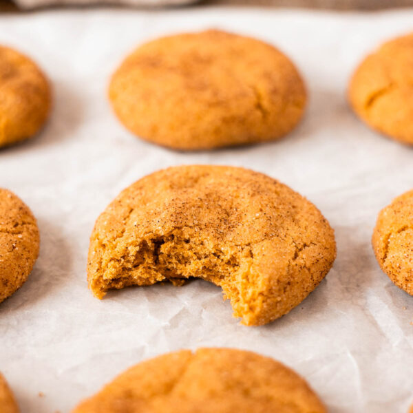 Several round pumpkin snickerdoodles are arranged on parchment paper, with one cookie in the center showing a bite taken out of it.
