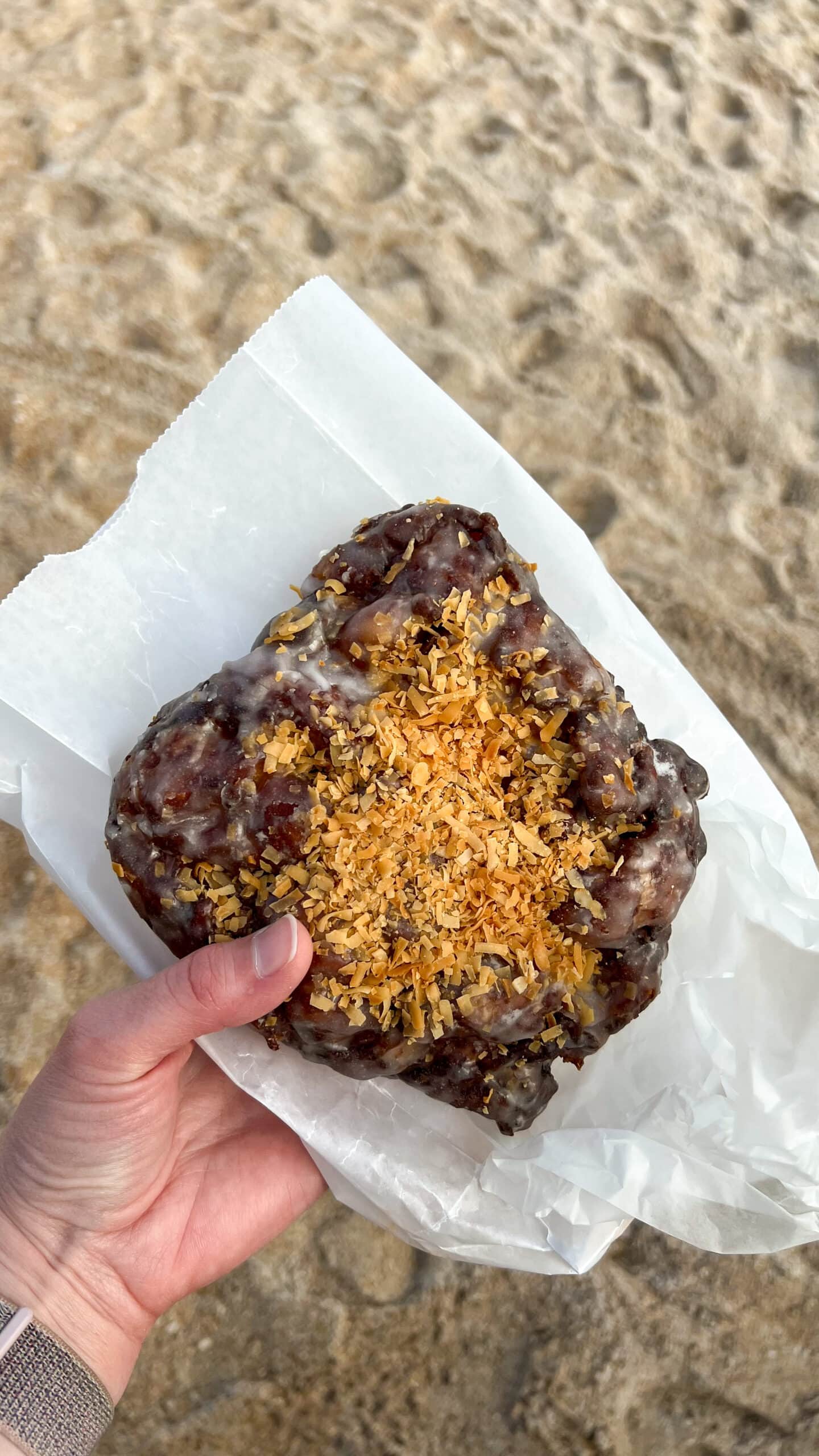 hand holding a large pineapple fritter topped with toasted coconut at the beach.