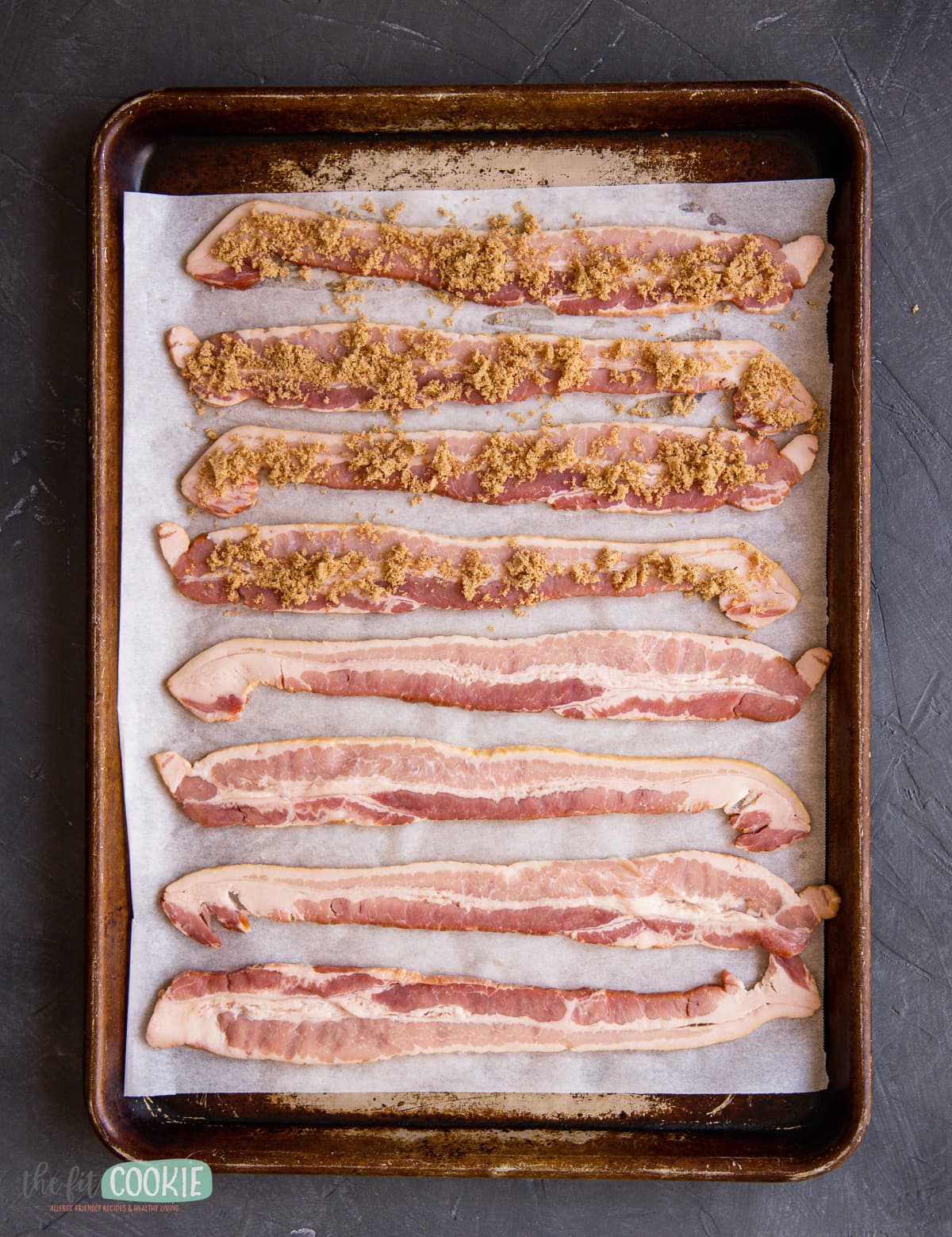 overhead photo of baking sheet pan with bacon on it, some of the bacon is covered with brown sugar. 
