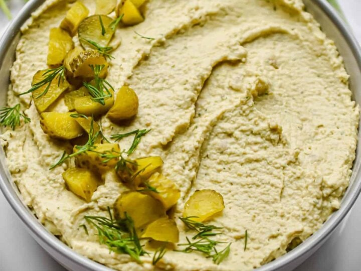 A bowl of hummus with pickles and dill on top.