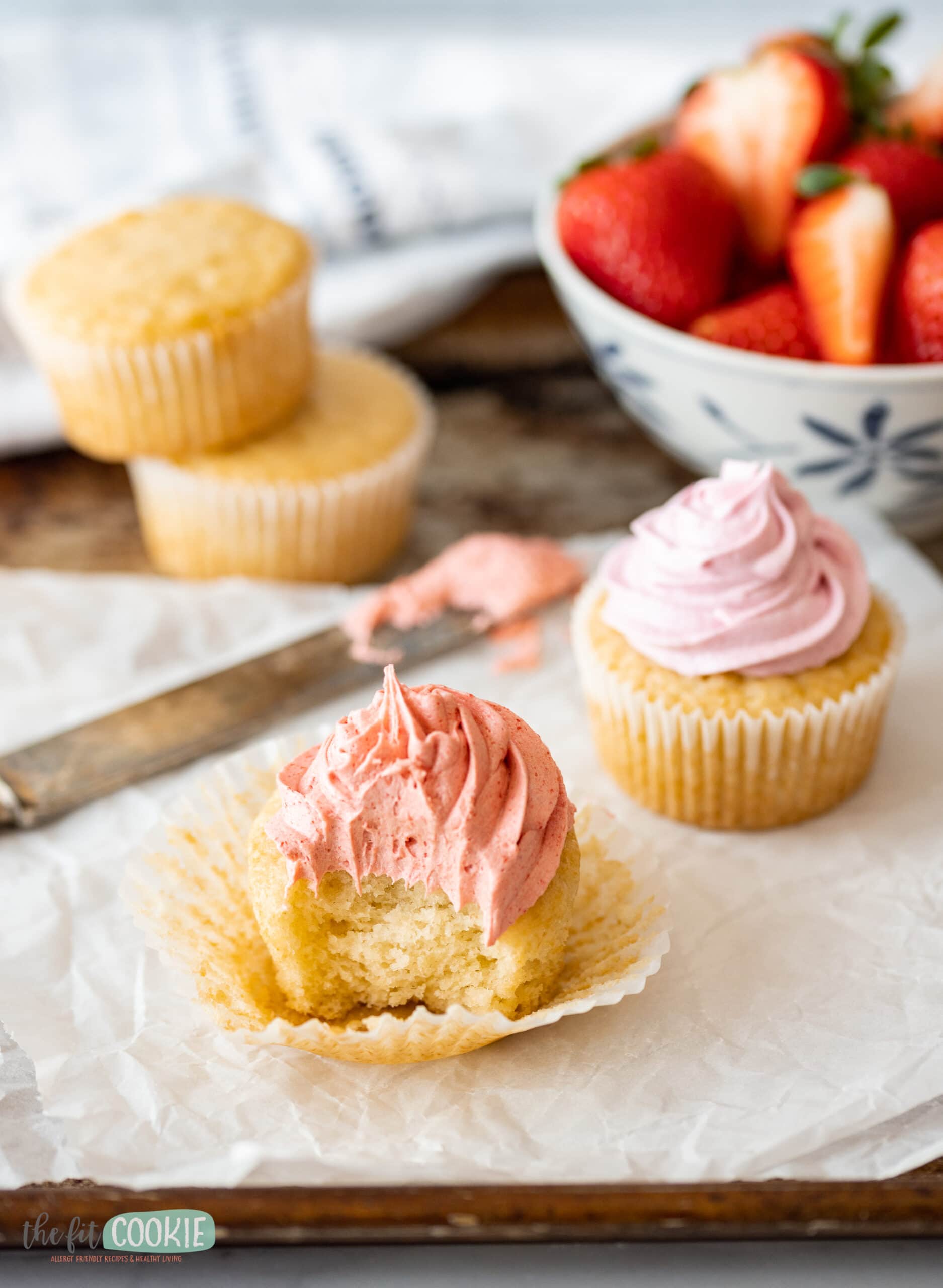 angled photo of vanilla cupcake with strawberry frosting and a bite taken. 