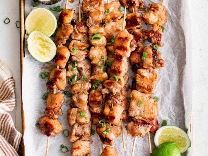 Chicken skewers on a baking sheet with lime wedges.