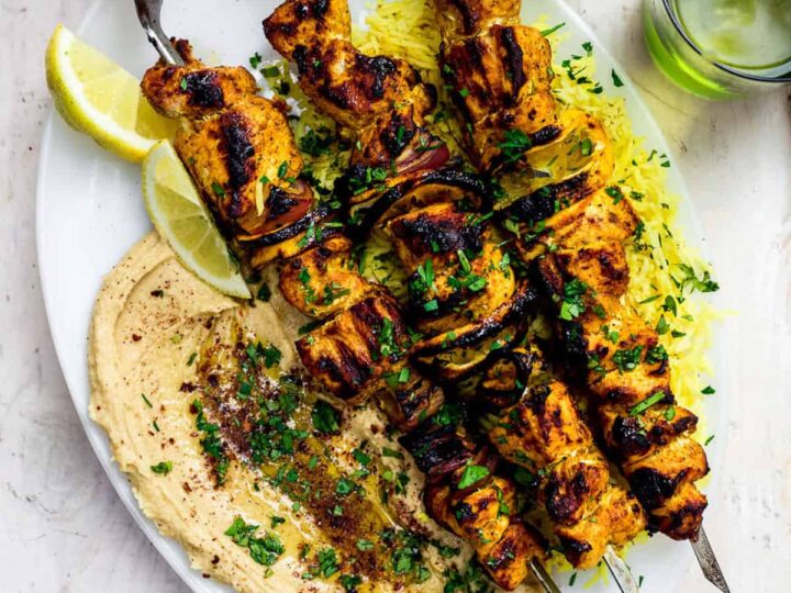 Chicken skewers with hummus and lemon wedges on a white plate.
