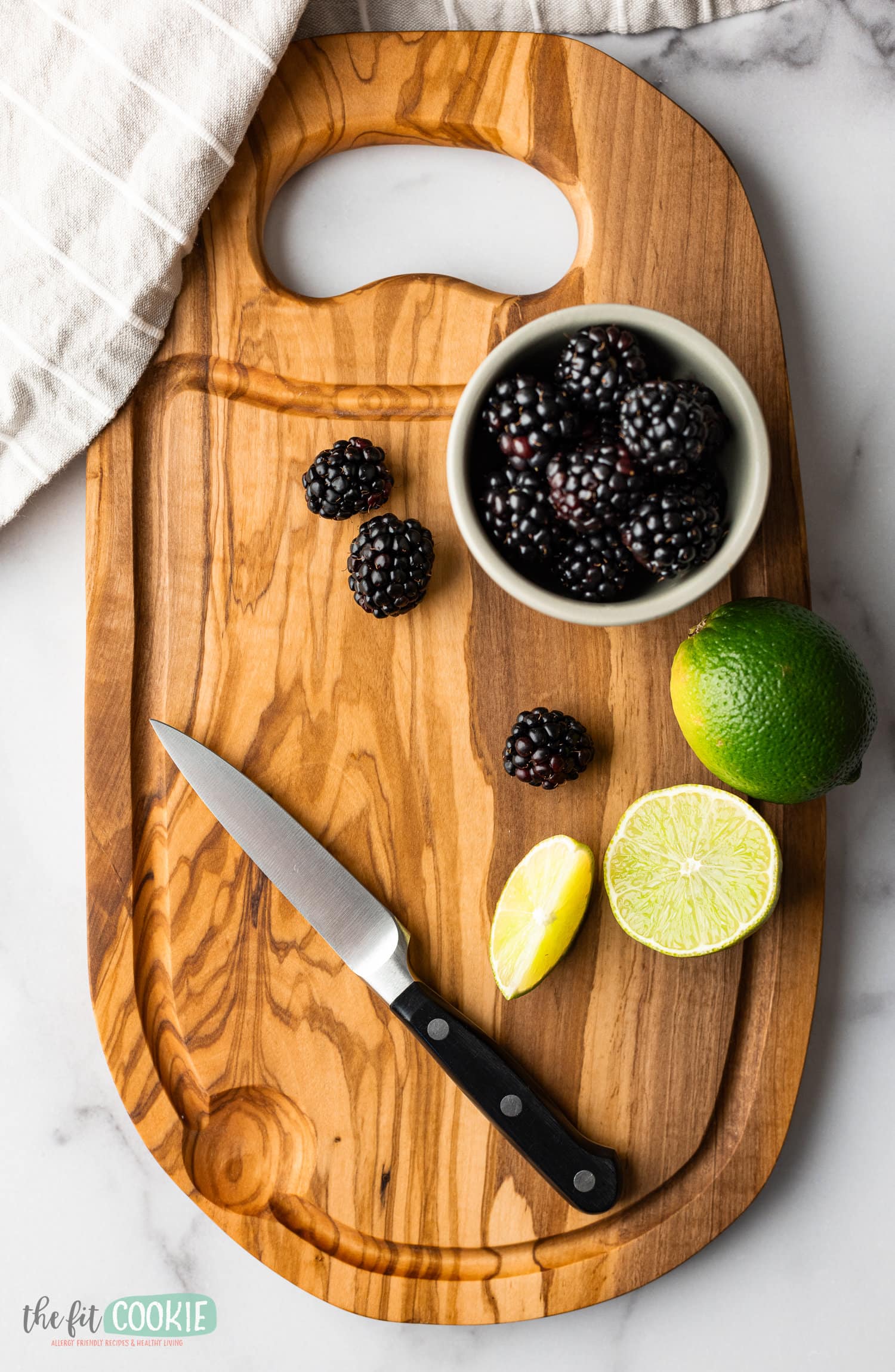 An olive wood cutting board with blackberries, limes, and a knife.