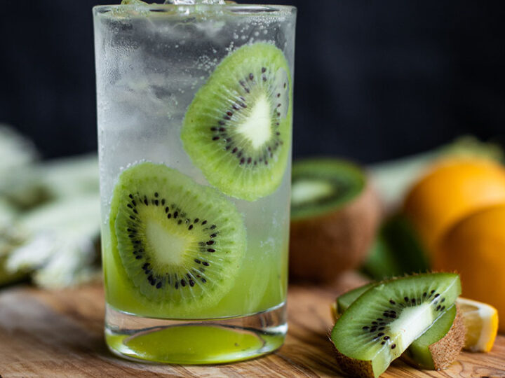 A drink with kiwi slices and oranges on a wooden cutting board.