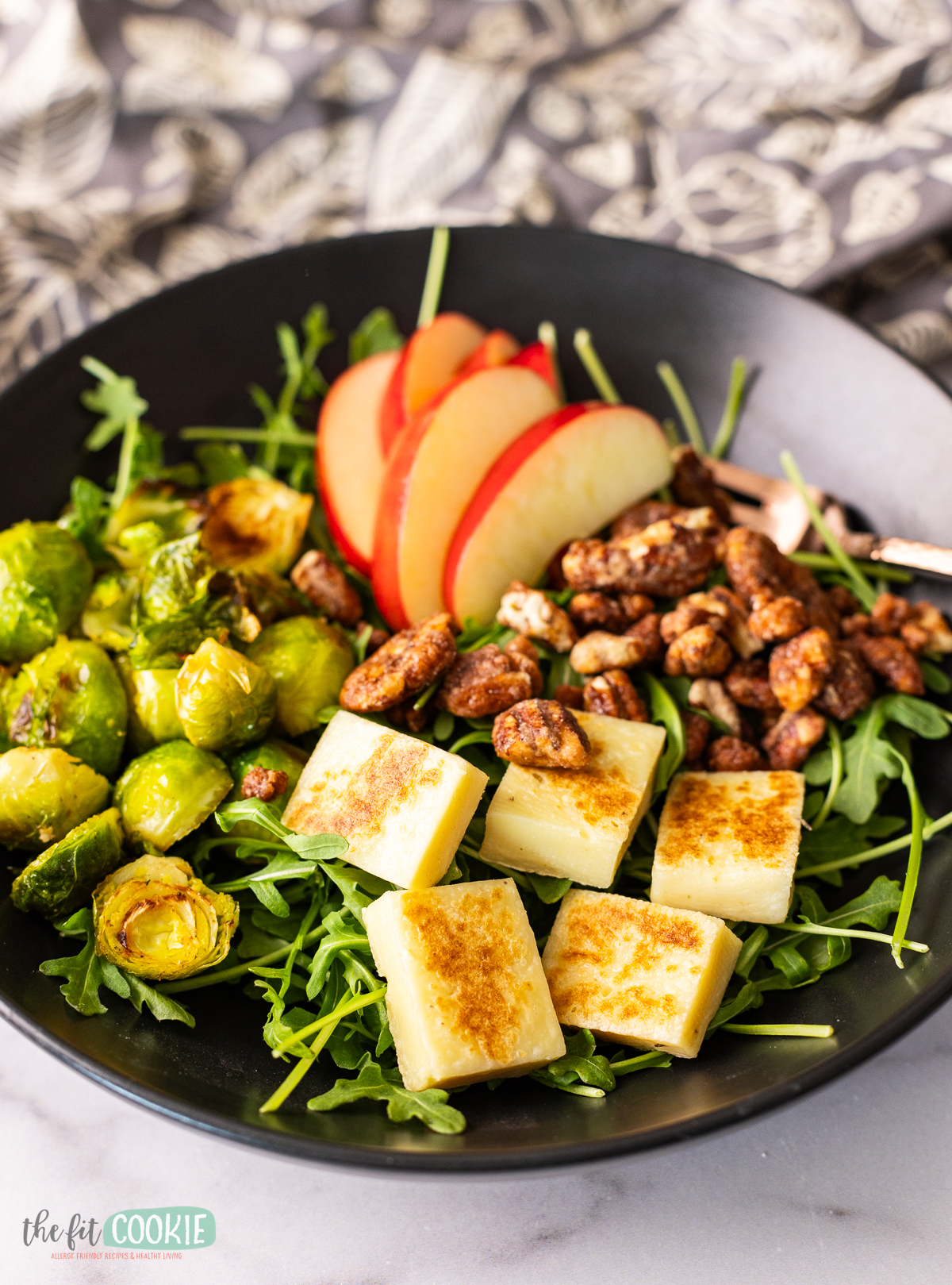 A winter salad in a black bowl filled with apples, brussel sprouts, and dairy free grilling cheese.