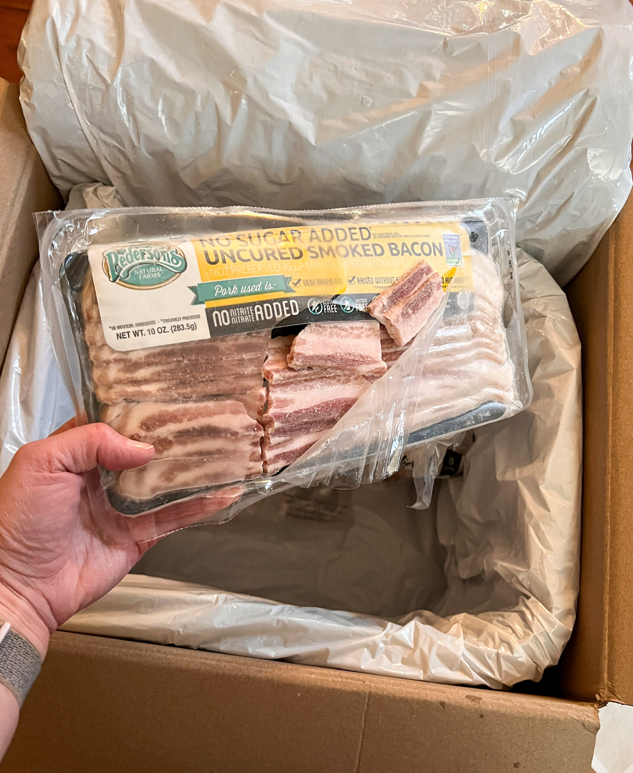 A person holding a package of broken bacon over a thrive market frozen box.