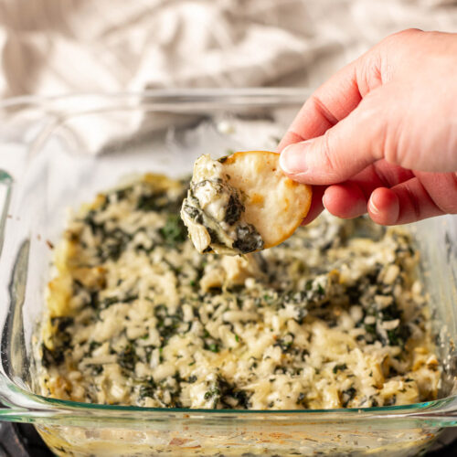 A person dipping a piece of bread into a dish of spinach artichoke dip.