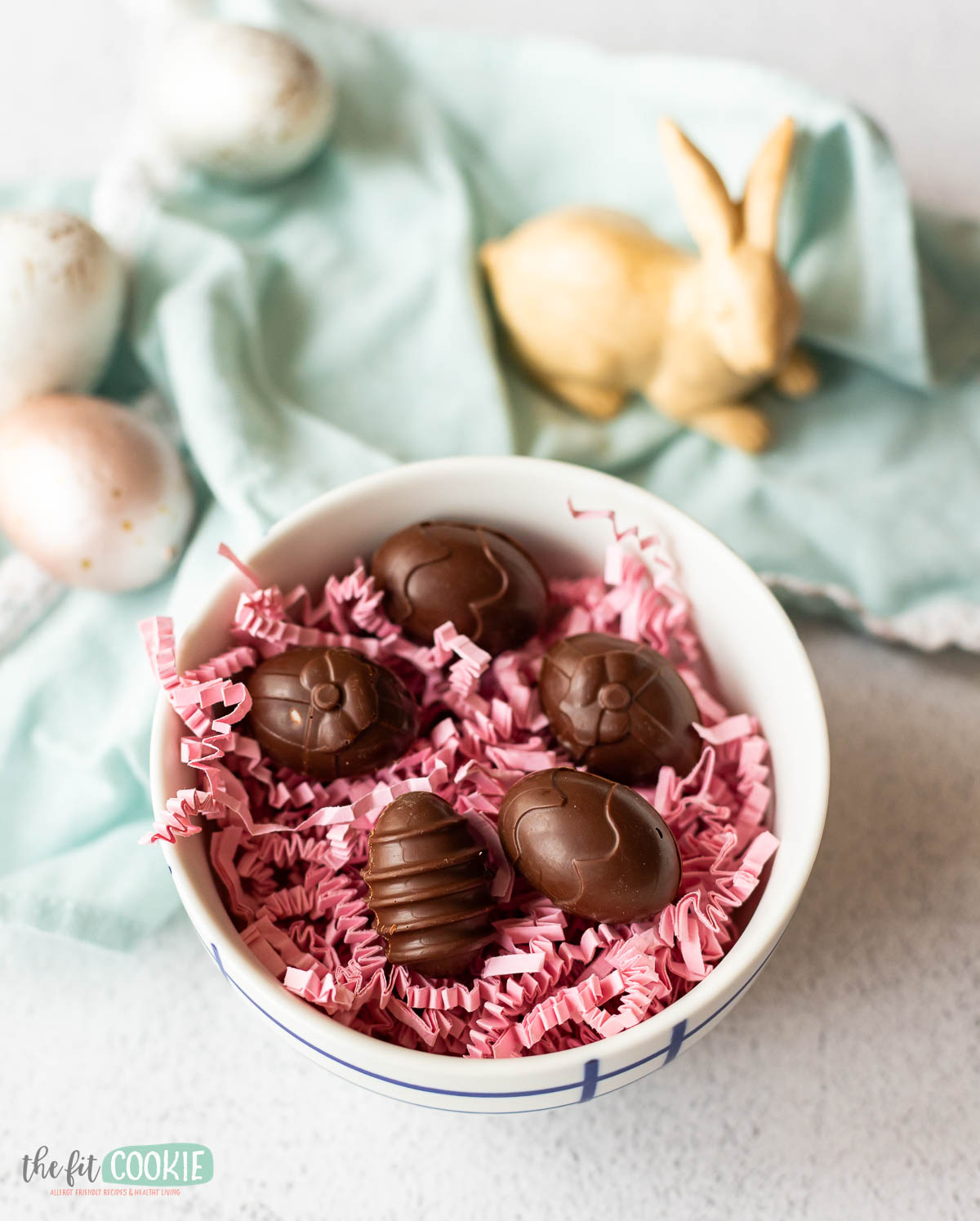 A bowl of chocolate cream eggs on a bed of pink shredded paper with a decorative rabbit and speckled eggs in the background.